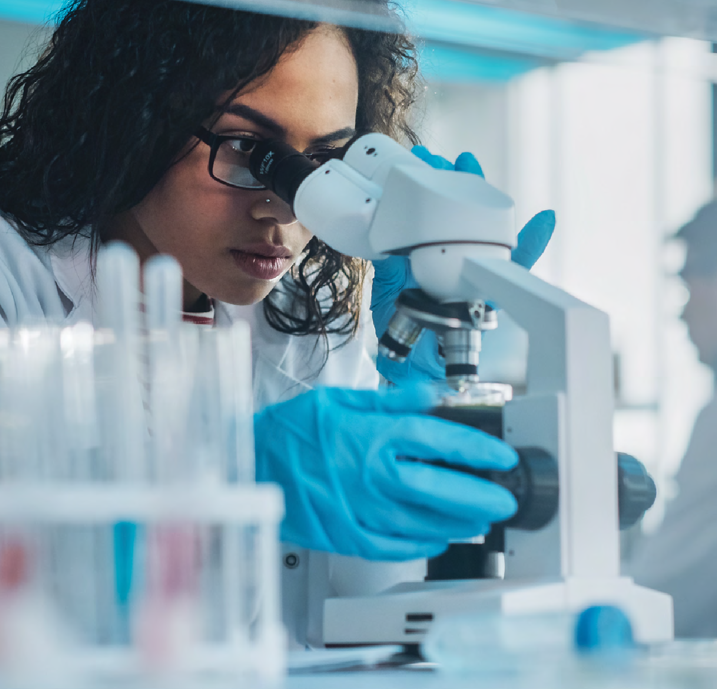 Laboratory technician wearing gloves examining a sample through a microscope in a medical lab setting.